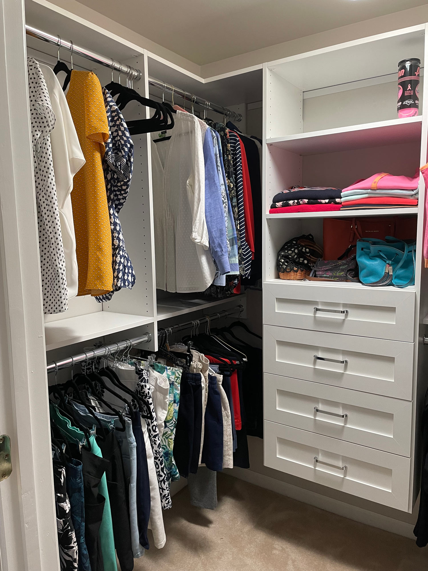 A neatly organized closet with hanging clothes, folded shirts, bags, and drawers on white shelves.