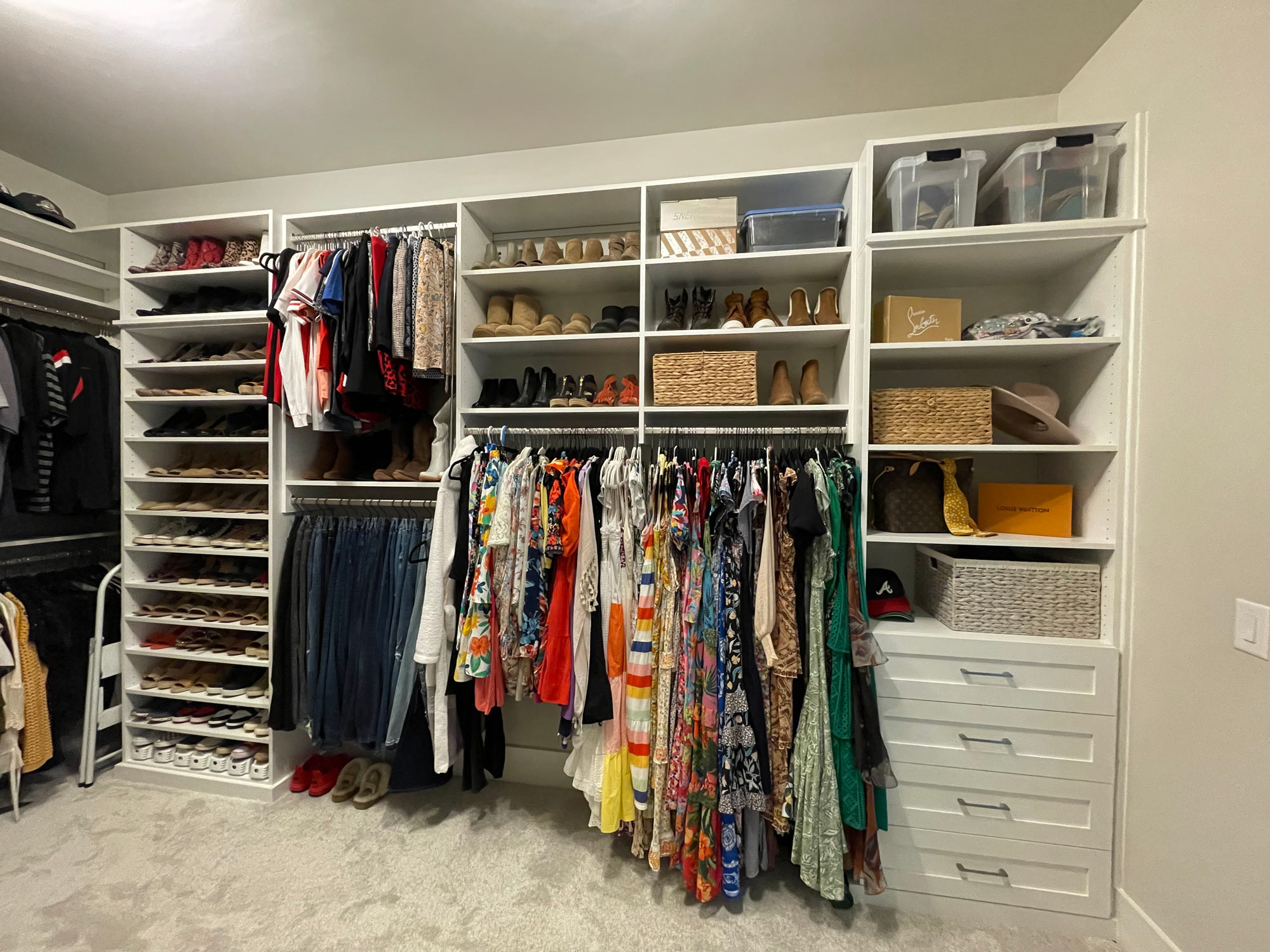 Organized closet with hanging clothes, shoes, storage bins, baskets, and shelves against a white wall.