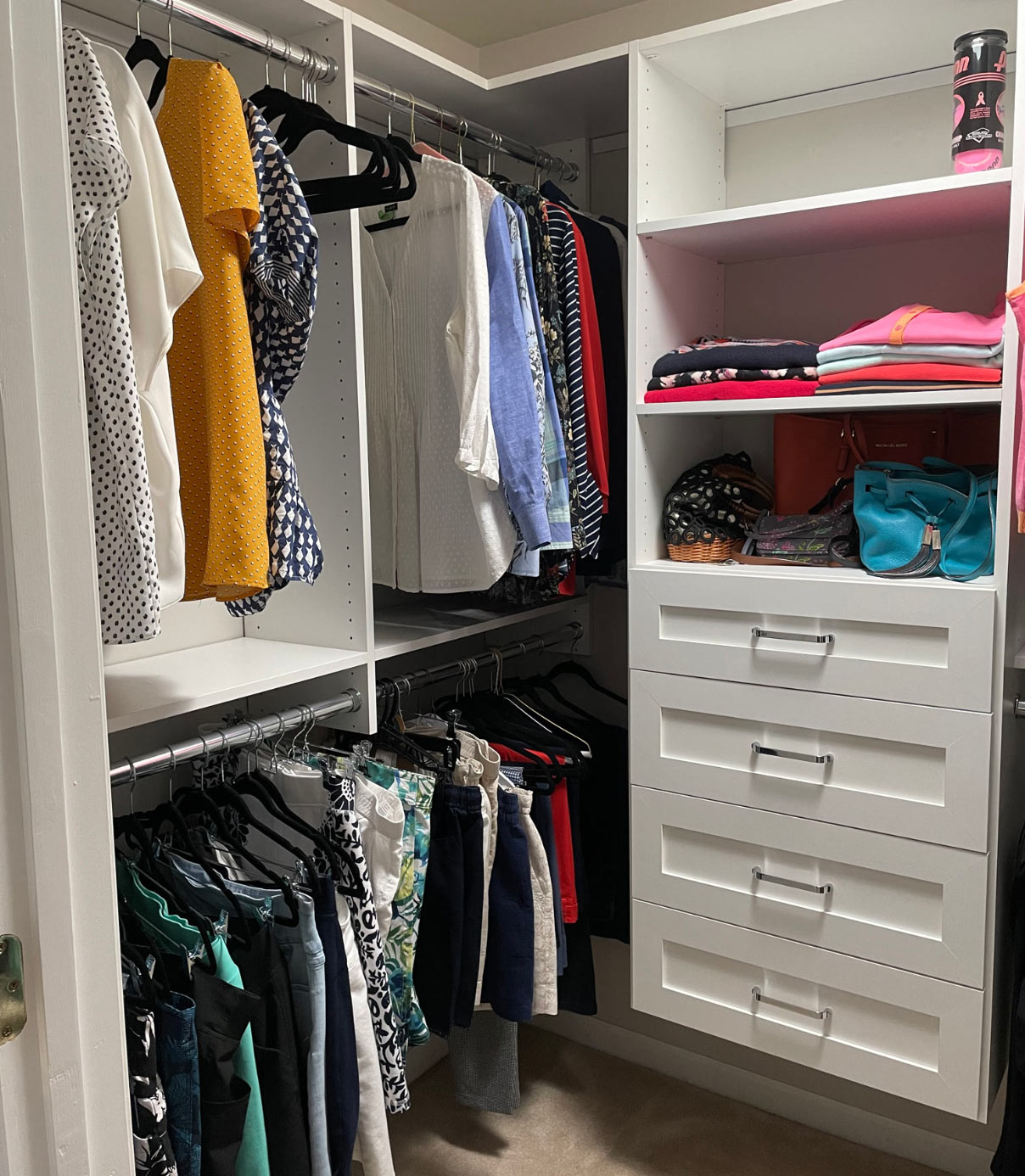 Neatly organized closet with hanging clothes, folded tops, bags, and drawers in a white shelving unit.