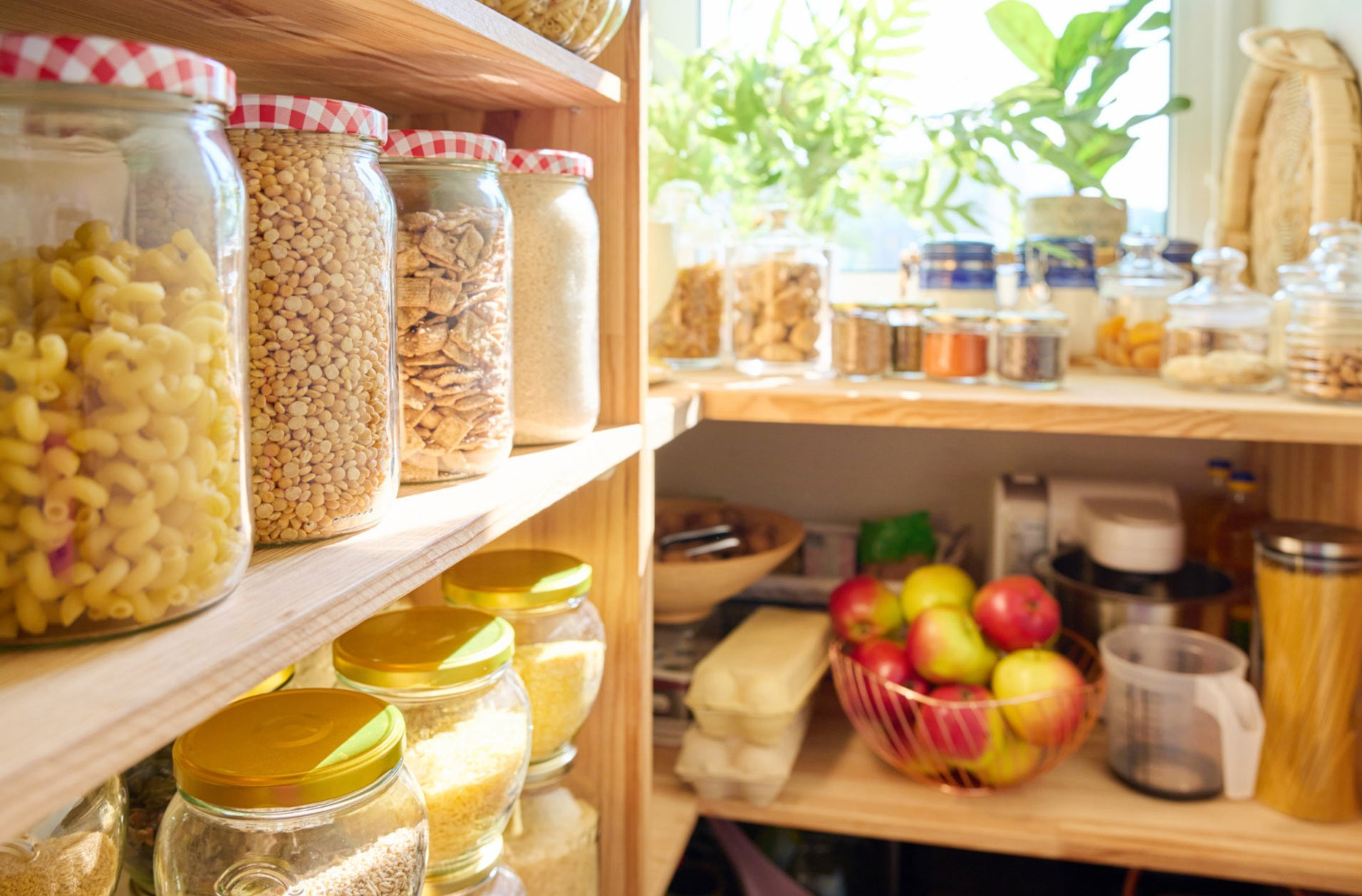 Glass jars of pasta, grains, and snacks on wooden shelves in a bright, organized pantry with fruit and plants.