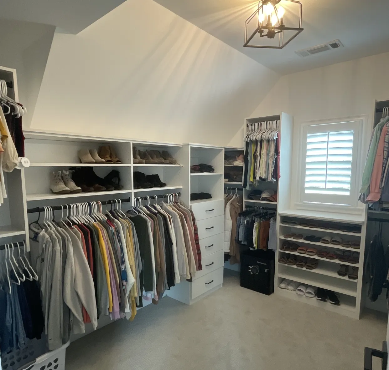 Walk-in closet with hanging clothes, shelves of shoes, drawers, and a window with blinds under a ceiling light.