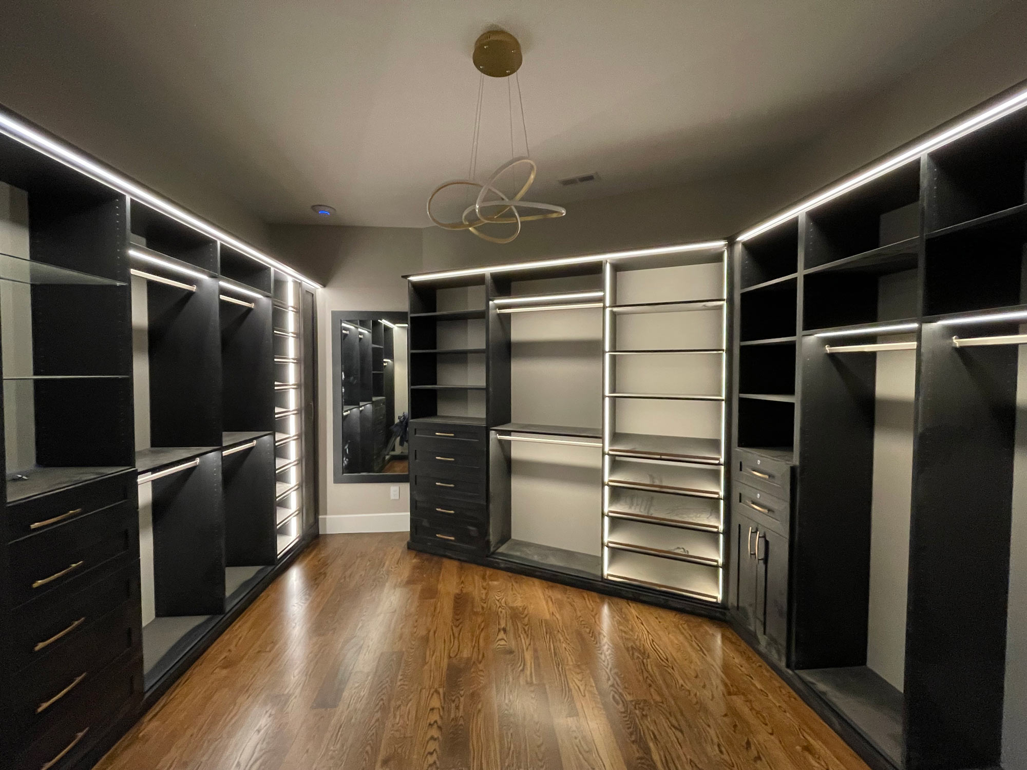 Primary-Walk-In-Black Modern walk-in closet with built-in shelves, drawers, hanging rods, and LED lighting on dark wood flooring.