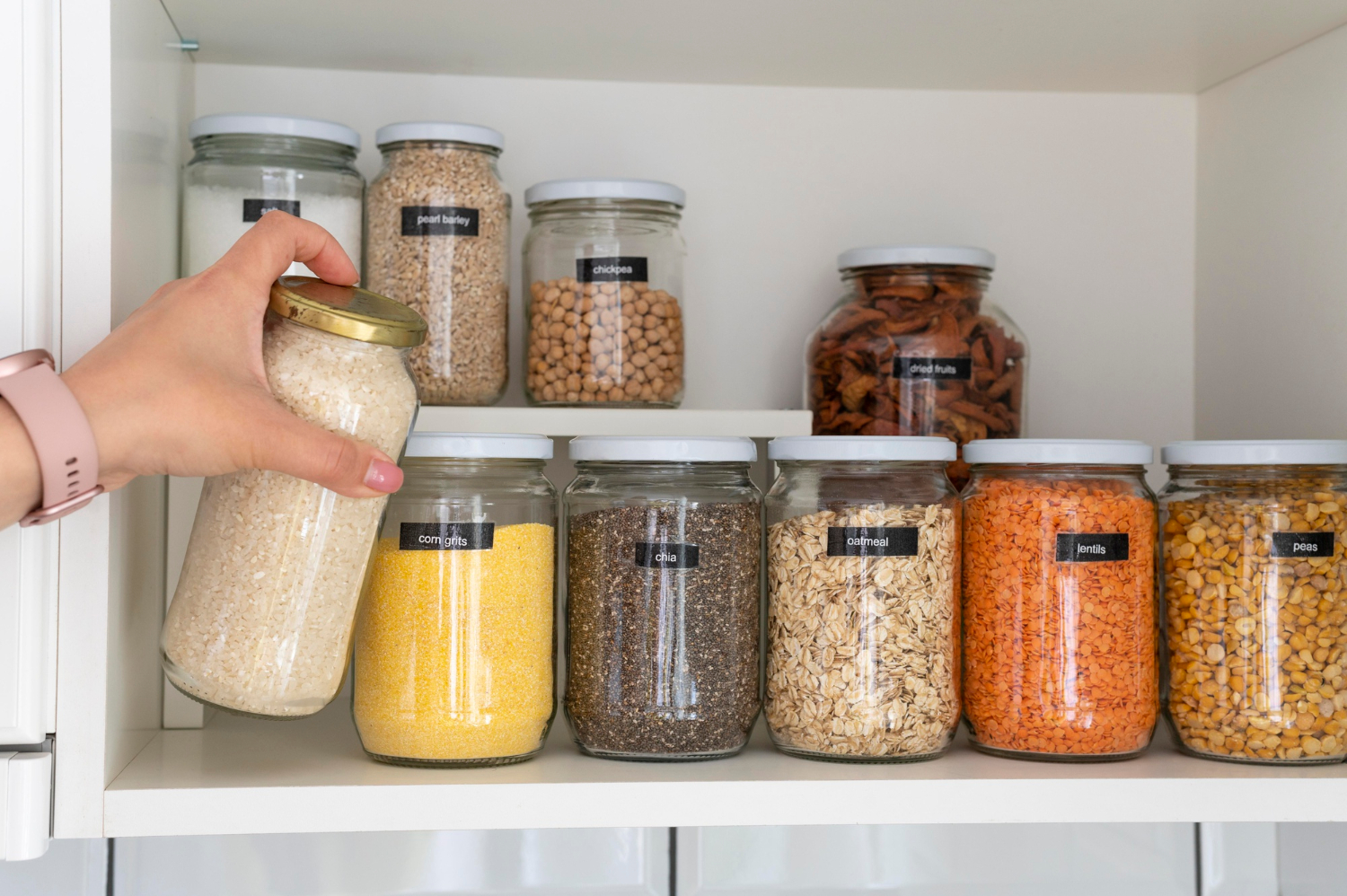 A hand takes a jar of rice from a shelf with various labeled jars of grains and legumes.